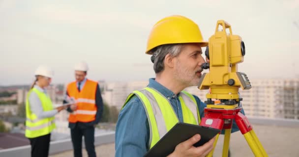 Kaukázusi férfi topográfus kaszkasztikus mérési szögben, teljes állomással az épület tetején. Férfi építtető építész topográfiai méréseket végez és táblagépet használ. Geodéziai fogalom. Eszközszámítógép. — Stock videók