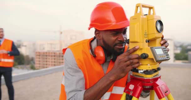 Afrikai amerikai fiatal férfi topográfus kaszkasztikus mérési szögben, teljes állomással az épület tetején. Férfi építész. Építész topográfiai méréseket végez. Geodéziai fogalom. Geodéziai. Építés. — Stock videók