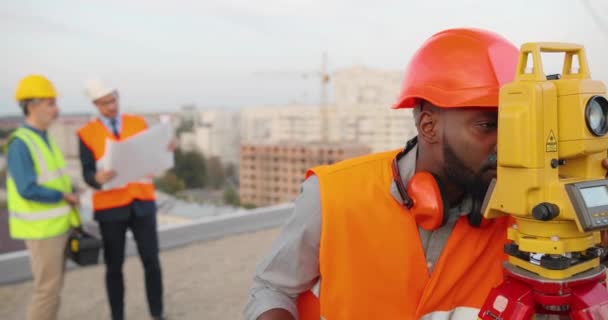 Afrikai amerikai férfi topográfus kaszkasztikus mérési szögben, teljes állomással az épület tetején. Férfi építtető építész topográfiai méréseket végez és táblagépet használ. Geodéziai fogalom. — Stock videók