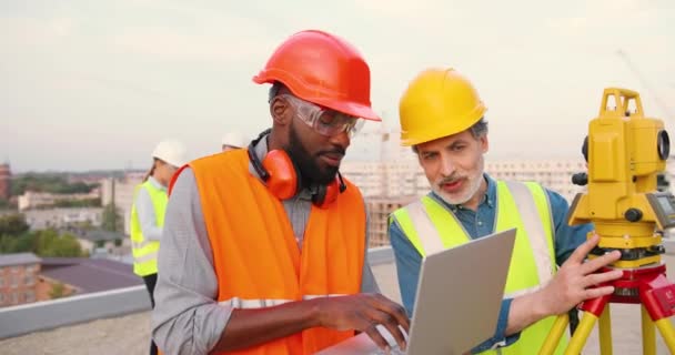 Carreras mixtas hombres constructores en el sitio de construcción de trabajo y ángulo de medición con la estación total en el techo. Topógrafos masculinos multiétnicos. Constructores caucásicos y afroamericanos con computadora portátil. — Vídeo de stock