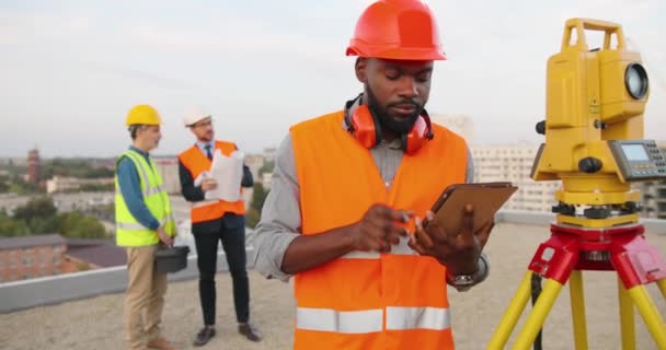 Αφροαμερικανός τοπογράφος σε casque γωνία μέτρησης με συνολικό σταθμό στην οροφή του κτιρίου. Αρσενικός κατασκευαστής οικοδόμος που κάνει τοπογραφικά μέτρα και χρησιμοποιεί συσκευή tablet. Γεωδαιτική έννοια Γεωδαιτική — Αρχείο Βίντεο