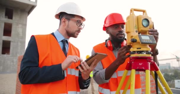 Szef rasy kaukaskiej w krawacie z tabletem i w kasku. Afroamerykanin męski budowniczy kąt pomiarowy z całkowitej stacji. Geodezja działa. Koncepcja topograficzna. Praca geodezyjna. — Wideo stockowe