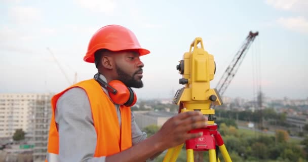 Porträt eines gutaussehenden jungen afroamerikanischen Mannes in Kaskade, der auf der Baustelle in die Kamera blickt und am Bahnhof lächelt. Gut aussehende männliche Bauarbeiter lächeln, wenn sie draußen bauen. Topograph. — Stockvideo