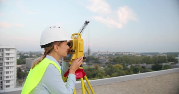 Topógrafo mulher bonita caucasiana em ângulo de medição em casco com estação total no telhado do edifício. Construtora feminina. Construtor fazendo medidas topográficas. Conceito de geodésia. Construção de obras. — Vídeo de Stock