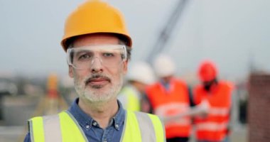 Şantiyede kameraya dönüp gülümseyen kasklı ve gözlüklü yakışıklı beyaz adamın portresi. Açık havada sakallı, yakışıklı bir erkek müteahhit. Konsept oluştur.