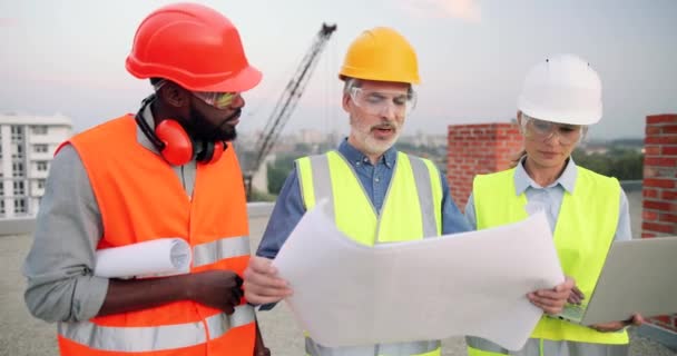 Kaukasische en Afro-Amerikaanse mannen en vrouwelijke bouwers, architecten en ingenieurs met ontwerp plan van de bouw en tablet praten op bouwplaats. Multi-etnische mannen en vrouwen constructeurs. — Stockvideo