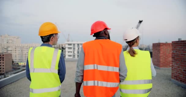 Achter. Gemengde-races mannelijke en vrouwelijke bouwers in casques lopen op het dak van de bouw en praten. Terugblik op multi-etnische voorman en werknemers die werken en discussiëren over de bouw. — Stockvideo