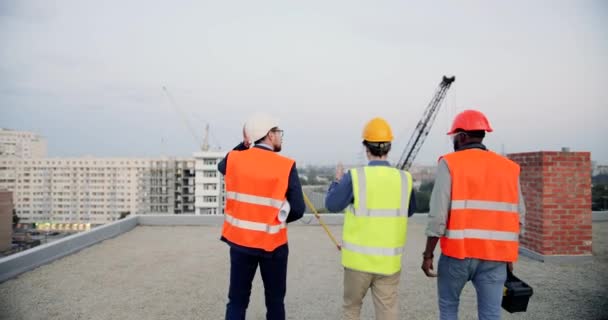 Achter. Senior wijze architect in bril en casque lopen op het dak van de bouw en praten met twee mannen bouwers. Terugblik op ploegbazen en arbeiders die werken en discussiëren over de bouw — Stockvideo