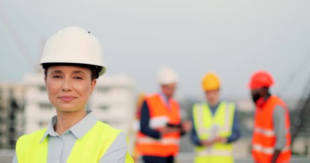 Retrato de la hermosa mujer joven caucásica en casco de pie en el sitio de construcción y sonriendo a la cámara. Constructora en el edificio. Multi hombres étnicos constructores que trabajan en el fondo. — Vídeo de stock