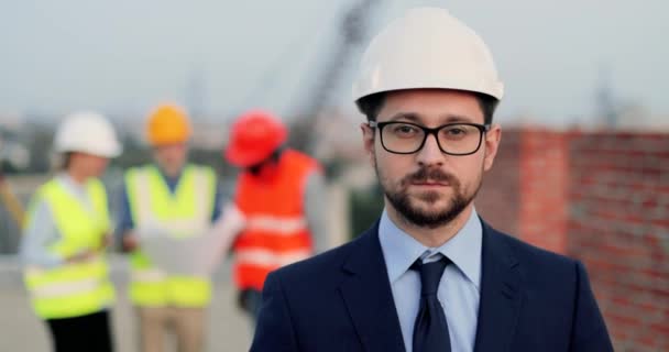 Portret van een knappe blanke jongeman architecte in casque, pak, das en bril op de bouwplaats die er recht uit ziet. Een mannelijke investeerder in de bouw. Gemengde-rassen constructeurs praten op de achtergrond. — Stockvideo