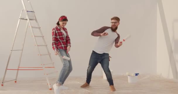 Feliz pareja joven haciendo reparaciones en un apartamento nuevo. Alegre hombre y mujer bailando con instrumentos sobre fondo de pared blanca y escalera. — Vídeos de Stock