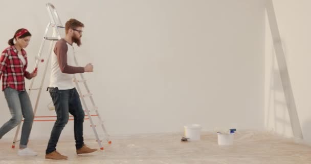Junges fröhliches Ehepaar tanzt in neuer Wohnung mit Werkzeug für Reparaturen in der Hand. Zwei Personen bewegen sich rhythmisch auf dem Hintergrund einer weißen Wand. — Stockvideo