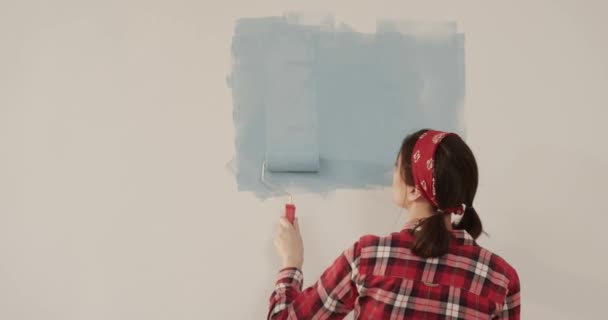 Joven hermosa mujer en camisa de cuadros rojos se encuentra en una habitación nueva y pinta la pared de color azul. La chica hace reparaciones por su cuenta. Vista desde atrás. — Vídeos de Stock