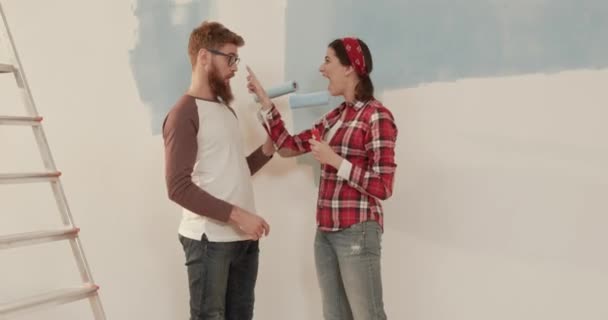 Una joven pareja de casados se mudó a un apartamento nuevo y haciendo reparaciones. Dos personas alegres se divierten sosteniendo rodillos de pintura. — Vídeos de Stock