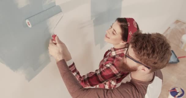 Feliz joven pareja casada pintando juntos la pared en azul sosteniendo un rodillo de pintura. El marido abrazó a su esposa. En cámara lenta. Vista superior. — Vídeos de Stock