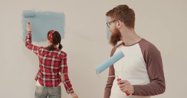 Porträt eines jungen, gut aussehenden Mannes, der mit einer Farbrolle in den Händen steht und lächelt, während eine Frau die Wand bemalt. — Stockvideo