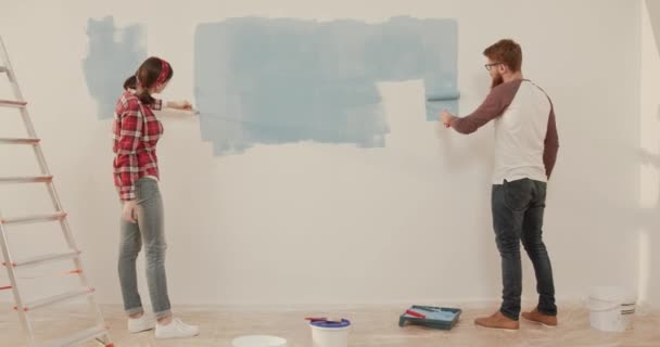 Pareja de jóvenes casados se centró en la pintura de la pared en azul en el nuevo apartamento. Dos personas hacen reparaciones en casa por su cuenta. — Vídeos de Stock