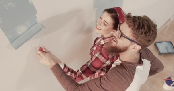 Mutlu, genç, evli bir çift ellerinde bir boya silindiri tutarak duvarları maviyle boyuyor. Koca, karısına duvar boyamasında yardım eder ve onu över.. — Stok video