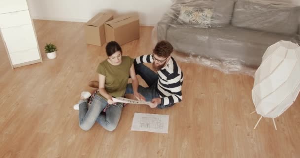 Νεαρό παντρεμένο ζευγάρι μετακόμισε σε νέο διαμέρισμα και κάθεται στο πάτωμα πάνω από το σχήμα χαρτί του διαμερίσματος σχεδιάζουν διάταξη των επίπλων. — Αρχείο Βίντεο