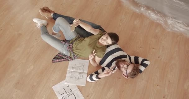 Unga glada gifta par ligger på golvet i en ny lägenhet och spela in videor på smartphone vinka åt kameran. Ovanifrån. — Stockvideo