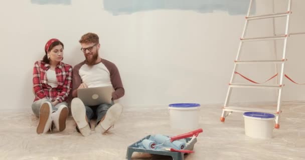 Unga gifta par sitter på golvet nära stege och hinkar med färg och titta på reparation tips på nätet på bärbar dator. — Stockvideo