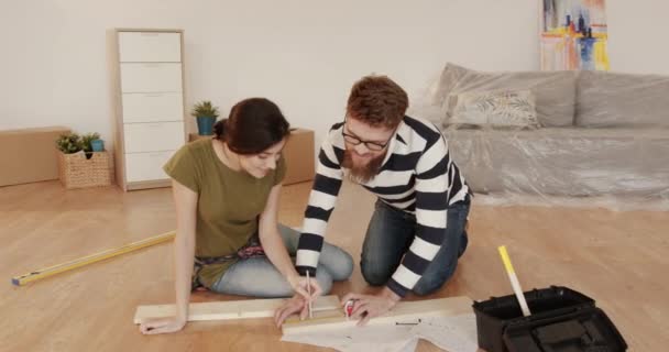 Young married couple sitting on floor in new apartment and assembling furniture together. Beautiful woman and man mark on wooden boards determining the appropriate size. — Stock Video