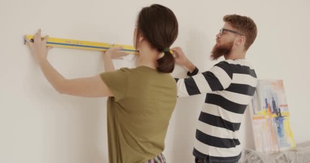 Junge Eheleute, die in der Nähe von Wandhalterungen stehen, um die Gleichheit der Oberflächen zu messen. Hübsch kaukasisch frau holding bau ebene mann help sie. — Stockvideo