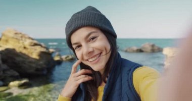 Güzel bir kızın, elinde kamerayla güldüğü ve poz verdiği portreyi yakından çek. Denizin arka planında selfie çekerken..