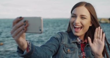 Mutlu genç kadın ve kot ceketin portresi uzanmış bir elle telefonu tutuyor ve deniz arka planında kameraya el sallıyor..