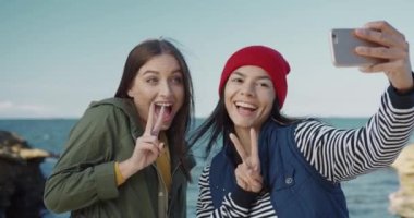 İki güzel beyaz kız denizin arka planında selfie çekiyor. Kadın arkadaşlar akıllı telefon kameraları için poz verir ve eğlenirler..