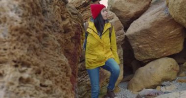Sarı ceketli güzel beyaz kadın turist deniz kıyısındaki taşların arasında yürüyor. Yalnız seyahat et. Kayaların ve kanyonların arasında yürü.