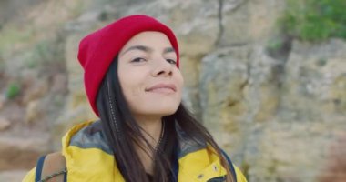 Kırmızı şapkalı, sırt çantalı, arka planda kayalara bakıp kameraya gülümseyen güzel kadın turistin portresini yakından çek..