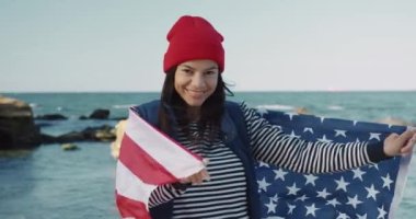 Genç, güzel, mutlu öğrenci kız kameraya döner ve Amerika bayrağına sarılmış bir şekilde gülümser. Değişim öğrencisi.