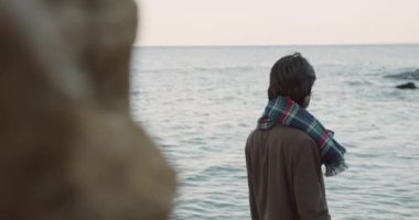 Yakışıklı, beyaz saçlı, şık ceketli, gözlüklü, kumsalda duran, başını çeviren ve kameraya bakan bir adam..