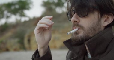 Gözlüklü beyaz adamın yakın portresi sigaraya üflüyor ve sahilde otururken saçlarını düzleştiriyor..