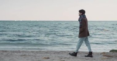 Ceketli ve atkılı yakışıklı genç adam ıssız sahilde yürüyor ve düşüncelere dalmış denize bakıyor..