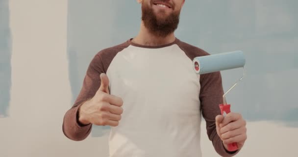 Primer plano de joven guapo hombre caucásico con barba sosteniendo rodillo de pintura y levantando el pulgar sonriendo. — Vídeos de Stock