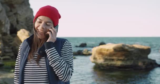 Ung vacker brunett kvinna i röd hatt talar på mobiltelefon stående på stranden nära stenar på sommaren. — Stockvideo