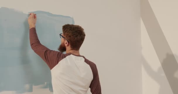 Junger Mann bemalt Wände zu Hause selbst mit Pinsel blau. Rückansicht. Konzept des Umzugs in neue Wohnung. — Stockvideo