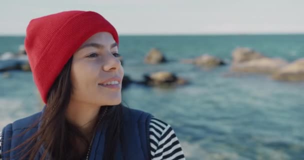 Close up retrato de mulher caucasiana atraente feliz em chapéu vermelho em pé junto ao mar e sorrindo olhando para a câmera. — Vídeo de Stock