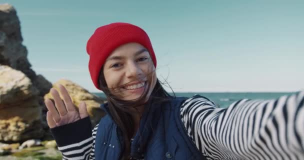 Close up of beautiful young happy woman waving hand at camera recording video message on sea background. — Stock Video