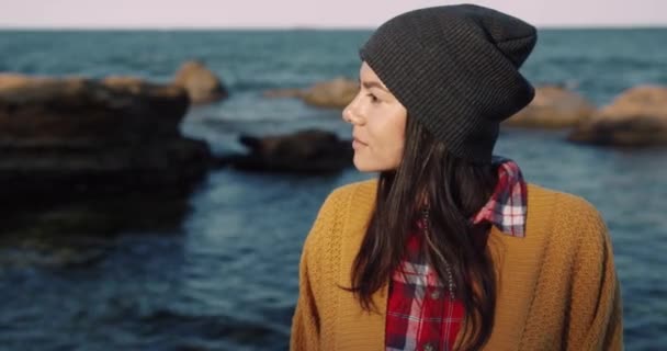 Portret van een gelukkige jonge vrouw met hoed en geel vest op de achtergrond van de zee en glimlachend kijkend naar de camera. — Stockvideo