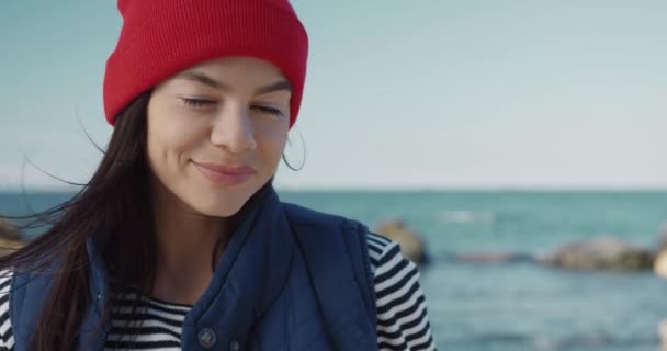 Primer plano retrato de mujer caucásica atractiva en sombrero rojo de pie en la costa y sonriendo mirando a la cámara. — Vídeos de Stock