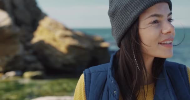 Ragazza felice in giubbotto blu e cappello guardando la fotocamera con bellissimi occhi marroni e sorrisi mentre in piedi contro il mare. — Video Stock