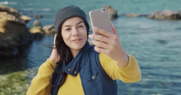 Portret szczęśliwej turystki w szarym kapeluszu trzymającej smartfona i rozmawiającej na wideo rozmowie machającej ręką stojącej nad morzem. — Wideo stockowe