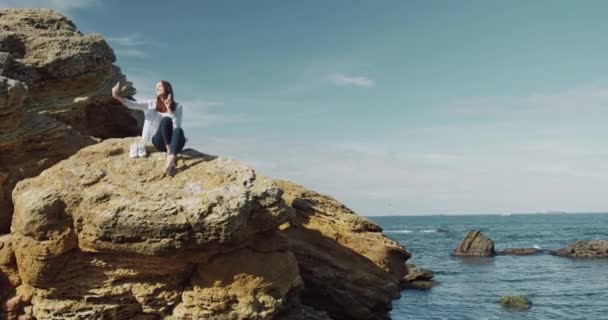 Kamera se pohybuje kolem krásné bělošky sedí na mořské skále, drží smartphone a natáčení videa na kameru. — Stock video