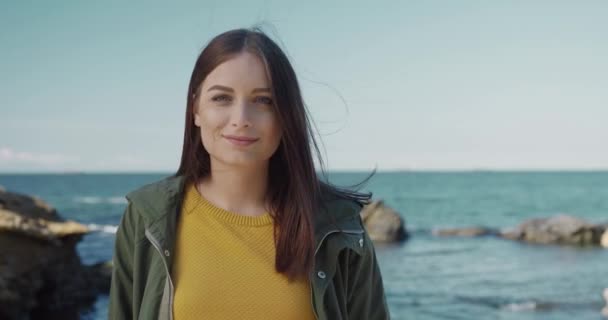 Retrato de bela menina caucasiana em jaqueta verde em pé no fundo do mar e pedras sorrindo enquanto o vento sopra seu cabelo. — Vídeo de Stock