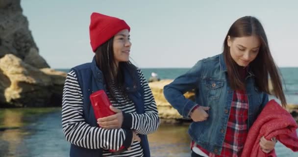 Duas amigas estão andando ao longo da costa do mar se divertindo conversando e segurando termo e cobertor para piquenique. — Vídeo de Stock