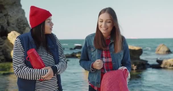 Dois melhores amigos estão andando ao longo da praia e conversando. Duas belas meninas caucasianas jovens estão andando ao longo da praia do mar com termo e cobertor nas mãos. — Vídeo de Stock