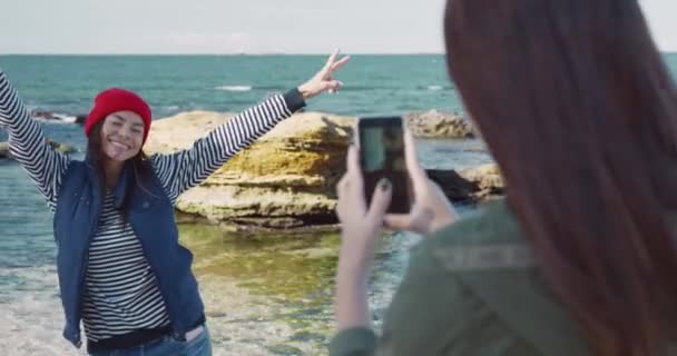 Vacker kaukasisk ung flicka i blå väst och röd hatt poserar på stranden för foto. Flicka tar bilder på sin vän på smartphone. — Stockvideo